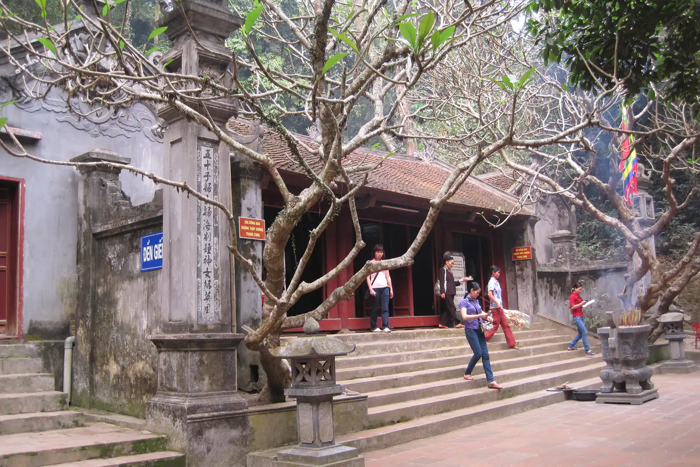 Đền Giếng trong khu di tích Đền Hùng, Phú Thọ: nếp đền mái ngói nâu, hàng cột gỗ sơn đỏ, bậc đá rộng dẫn xuống sân gạch; vài người đi lễ đang bước xuống bậc, bên trái là cột trụ đá khắc chữ Hán và tấm biển xanh “Đền Giếng”, phía trước có đèn đá và lư hương, cành cây đại không lá vắt ngang ảnh.