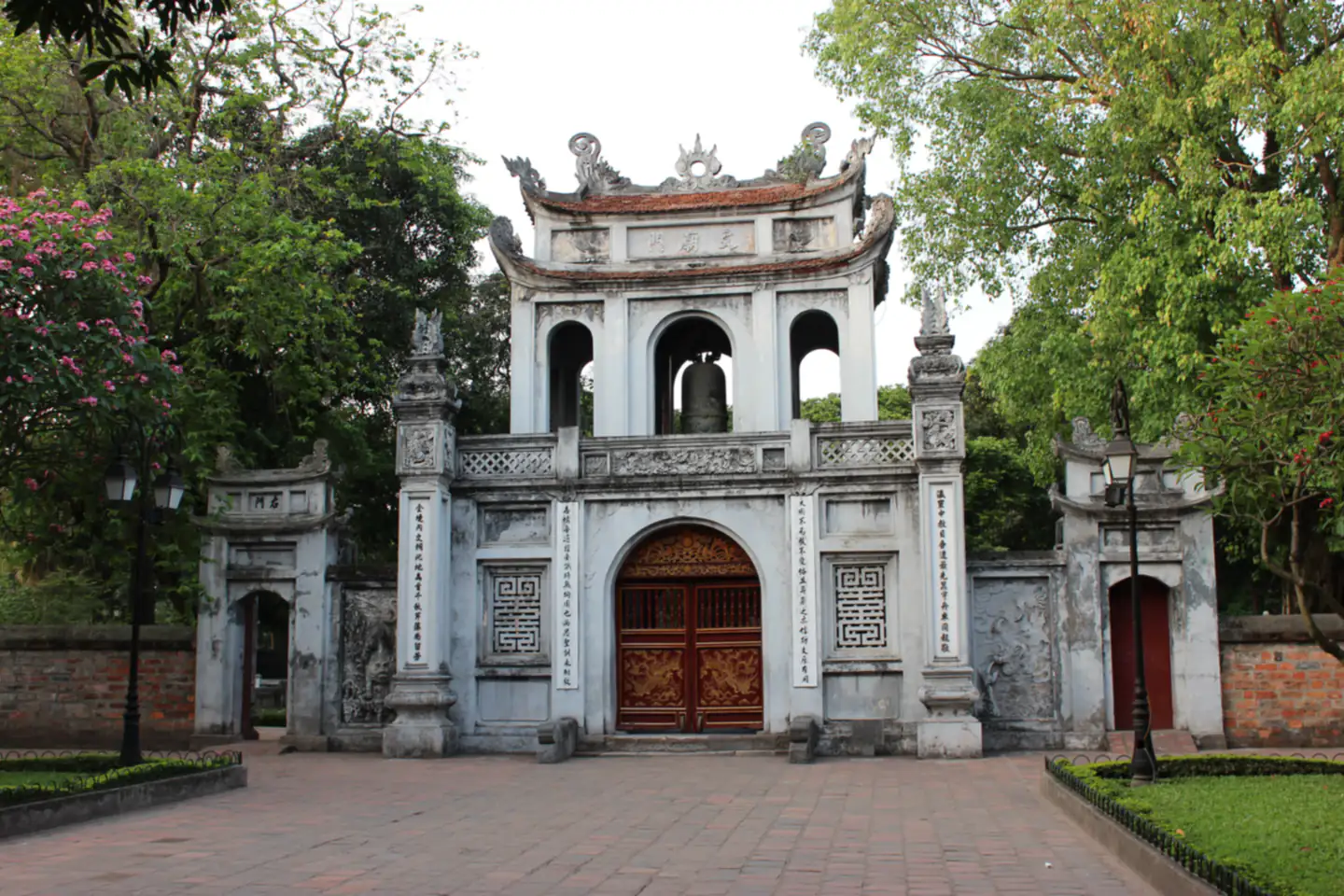 Cổng Văn Miếu Môn ở Văn Miếu – Quốc Tử Giám, Hà Nội: cổng tam quan tường trắng, gác trên có quả chuông đồng treo giữa ba ô vòm, hai cột trụ khắc câu đối đứng hai bên cánh cửa gỗ chạm rồng, xung quanh là cây xanh và sân gạch.
