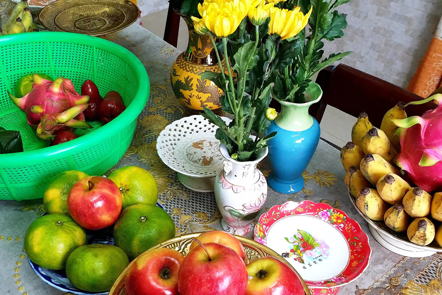 Góc bàn thờ ngày Tết của một gia đình Việt: đĩa táo đỏ, đĩa cam sành, nải chuối, quả thanh long và rổ roi bày trên khăn trải bàn thêu hoa, giữa những bình hoa cúc vàng, bình gốm xanh và đĩa sứ vẽ hoa.
